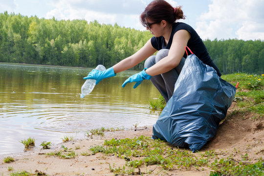 Image of woman in rubber gloves with dirty plastic bottle in her hands on river bank