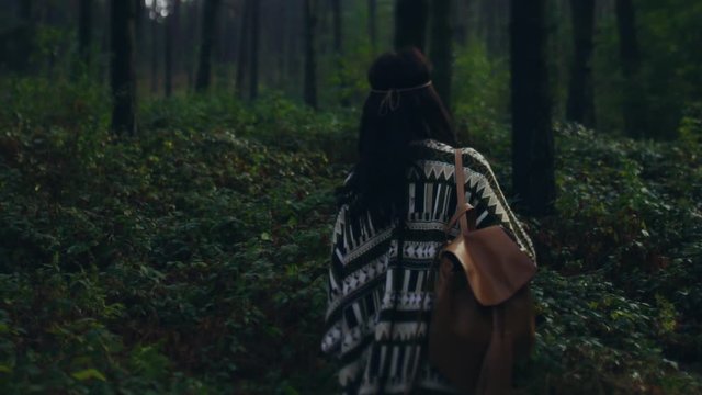 Back view of stylish attractive hippie girl walking through the dark forest. Active lifestyle, loneliness, wandering. Adventure time concept, travelling on foot. Follow me