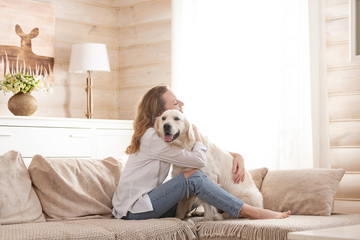Young pretty woman in casual clothes hugging her beloved big white dog sitting on the sofa in the living room of her cozy country house. Animal communication concept