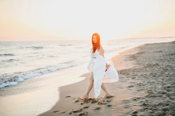 young beautiful girl in a white dress walks on the beach near the sea at sunset