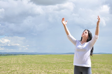 a woman stands with her arms raised and prays to God. On a green field in summer. Blue sky and white clouds.