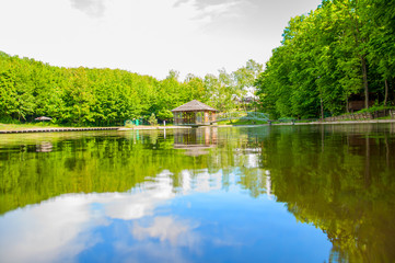 Fototapeta na wymiar In the summer on the lake. Wooden house on the shore of the pond. Reflection of blue sky clouds in water. Green trees.