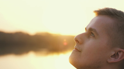 Close up portrait of pensive handsome young man looking up enjoying nature in sun rays at sunset.