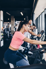 Young and attractive woman and man biking in the fitness gym. They exercising legs and doing cardio workout while riding cycling machines.