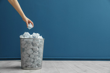 Woman throwing crumpled paper into metal bin against color wall. Space for text