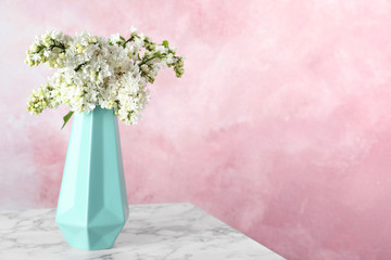 Blossoming lilac flowers in vase on marble table against color background. Space for text