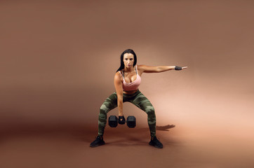 Fototapeta na wymiar Beautiful brunette hair fitness model wearing pink sports bra and camouflage tights holding a dumbbell in a squat position against a brown background