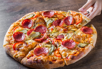 female hand takes a slice of Pepperoni Pizza with Mozzarella cheese, salami, bacon, Tomato sauce, pepper, Spices and pickled cucumbers. Italian pizza on wooden table background