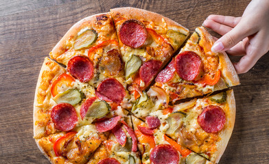female hand takes a slice of Pepperoni Pizza with Mozzarella cheese, salami, bacon, Tomato sauce, pepper, Spices and pickled cucumbers. Italian pizza on wooden table background