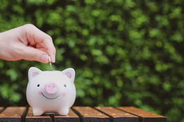 Man hand putting a coin to save in the white piggy bank with green background. Saving money for...