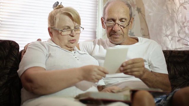 an adult couple looks at their photos sitting at home on the couch. husband and wife with glasses watching his album.