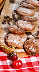 Oatmeal chocolate cookies with coffee grains and cherry, powdered sugar on kitchen cutting board with checkered fabric. Baking will charge you with energy and lift your spirits.