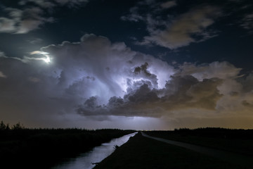 A majestic storm cloud is illuminated by lightning while the moon visible to the left