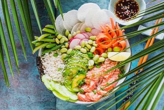 Poke bowl with salmon, avocado, edamame beans, vegetables and rice. Turquoise background and palm leaves sun light top view. 