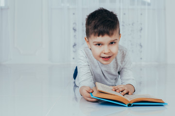 Closeup portrait of charming boy lying on the floor, smiling happily, reading a book. The boy is looking at the camera.