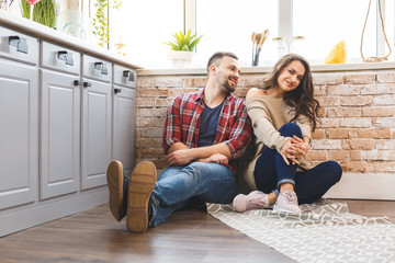 Young man and woman sitting on floor in kitchen and talking. Loving young couple spending time together at home.