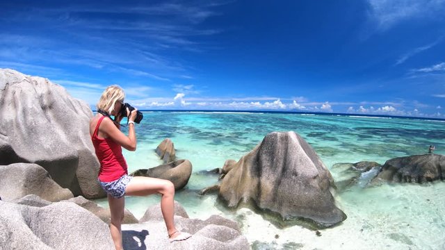 Lifestyle blonde woman photographer with camera at Anse Source d'Argent, La Digue, Seychelles. Shallow waters, pristine sand, turquoise sea and shaped boulders. Summer tropical holiday destination.