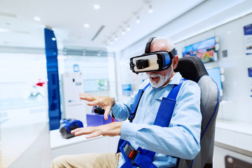 Excited bearded senior man sitting in chair and trying out virtual reality goggles. Tech store interior.