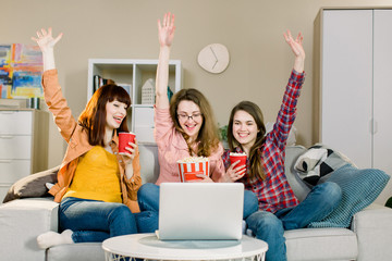 Happy funny crazy young female friends eating popcorn on sofa at home, while watching sport match and holding arms up