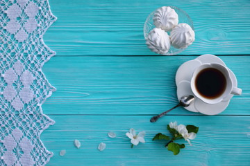A white cup of tea with a saucer and a spoon, a dessert of marshmallow, apple flowers and petals, white lace on a blue wooden table, top view
