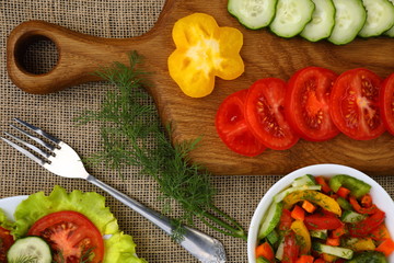 A cutting board with slices of tomatoes, cucumbers and pepper, a fork and two plates of vegetable salads, a branch of dill, healthy food on sack cloth as a background, horizontal, top view