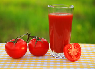 A glass of tomato juice with tomatoes on a yellow-and-white checkered tablecloth, blurred green natural background 