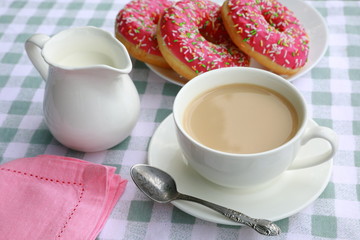 Breakfast composition of a cup of tea with cream, a creamer, a plate with pink donuts on a green-and-white checkered tablecloth 