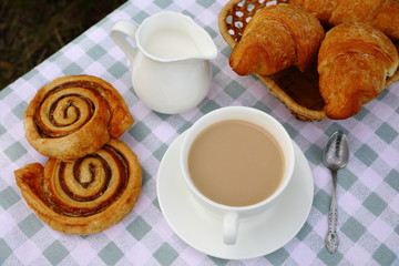 Breakfast composition of a cup of tea with cream, a creamer, bakery and croissants on a green-and-white checkered tablecloth, blurred green natural background