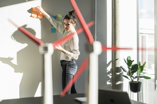 Businesswoman in office putting sticky notes on wall with wind turbine models on desk