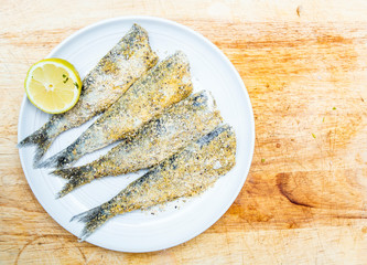 fresh sardines on a white plate with lemon on a wooden chopping board, seasoned with salt, pepper, oil, polenta and flour