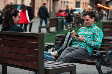 Fototapeta na wymiar laughing talking man with woman sitting on bench drinking coffee surfing in internet. urban lifestyle