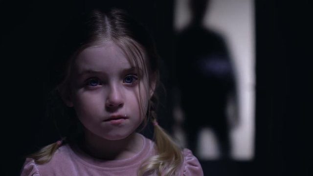 Little crying girl looking at camera, silhouette of rude father outside door