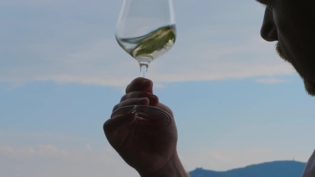 Man swirling, sniffing, tasting white wine in wineglass against blue sky. Person enjoying scent of wine, drinking on vacation. Glass with alcohol in bewhiskered winemaker hand in restaurant. Sommelier