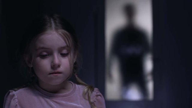 Crying little kidnapped victim looking at camera, criminal shadow outside door