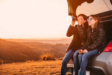 Mother and daughter sitting in a car trunk and using binoculars