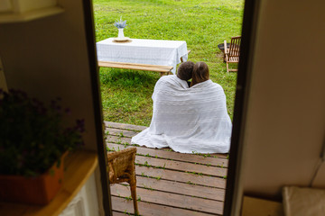 View from the door of the camper to a happy couple wrapped in a knitted blanket sitting next to the camper and warm up. Travelers sit on the wooden floor next to the house on wheels.