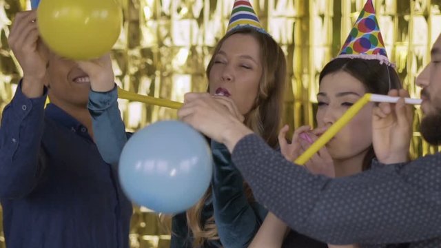 4 happy cheerful people at a party in paper caps fighting balloons. The guys are blowing in the pipes and dancing. A group of laughing people of mixed race on a gold background. HD