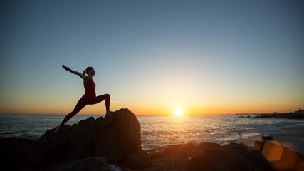 Silhouette young yoga woman doing fitness exercises on the sea beach at amazing sunset.
