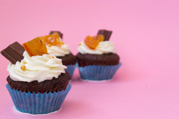 Three delicious cupcakes on a pink background. White air cream cake decorated with chocolate and candied fruit. Cupcakes in blue paper cups. There is a place for text.