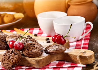 Oatmeal chocolate cookies with coffee grains and cherry, powdered sugar on kitchen cutting board with checkered fabric on wooden table in village style for picnic. Sunlight in kitchen.