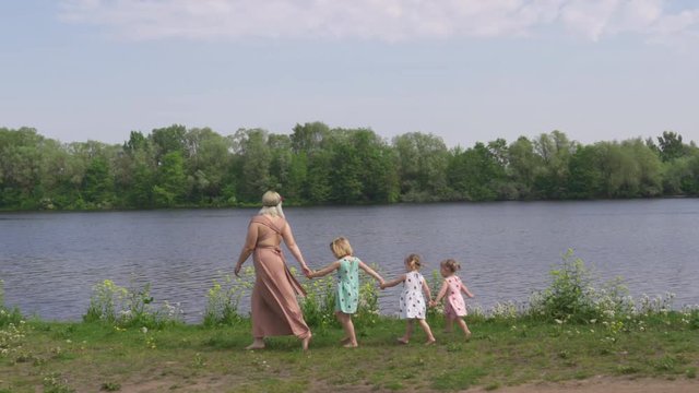 Young blonde hippie mother having quality time with her baby girls at a park - Daughters wear similar dresses with strawberry print - Walking like geese in a row along the river