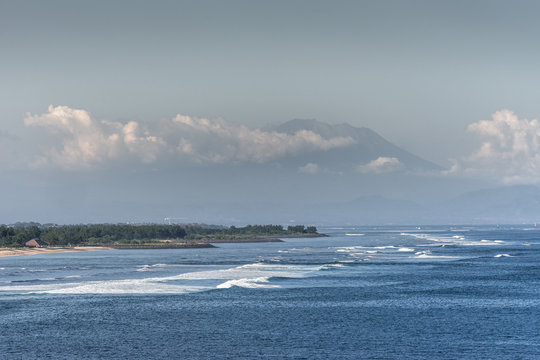 Bali, Indonesia - February 25, 2019: Ji Pantai Serangan beach north of Benoa Harbour Entrance. Blue water, sandy beach, green forest belt, tall mountain partly hidden in white clouds over light blue s