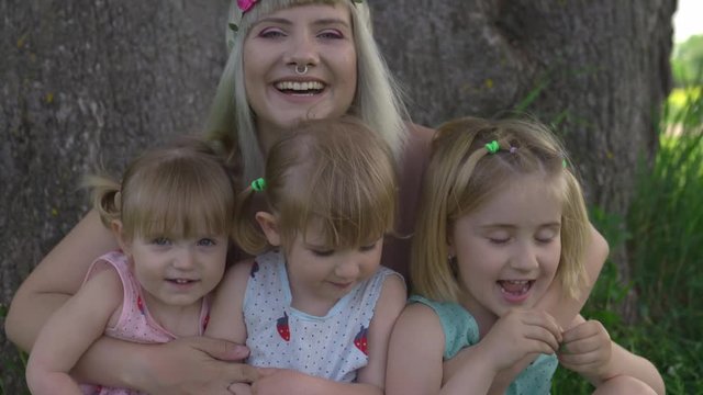 Young blonde hippie mother having quality time with her baby girls sitting and laughing under a tree in a park - Daughters wear similar dresses with strawberry print - Family values