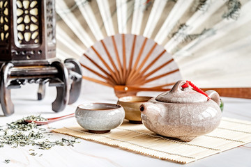 Asian teapot with teacups on bamboo tablamat decorated with chinese fan, lantern and scattered green tea on white marble background