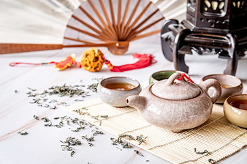 Asian teapot with teacups on bamboo tablamat decorated with chinese fan, lantern and scattered green tea on white marble background