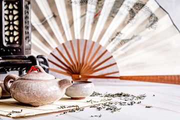 Asian teapot with teacups on bamboo tablamat decorated with chinese fan, lantern and scattered green tea on white marble background