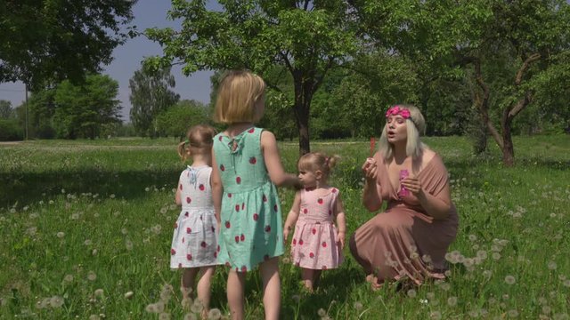 Young blonde hippie mother having quality time with her baby girls at a park blowing soap bubbles - Daughters wear similar dresses with strawberry print - Family values concept