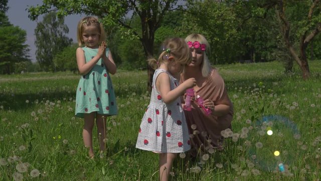 Young blonde hippie mother having quality time with her baby girls at a park blowing soap bubbles - Daughters wear similar dresses with strawberry print - Family values concept