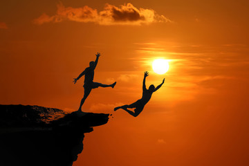 The concept of betrayal and the help of friends, Silhouette of Men are jumped between high cliff at a red sky sunset - obrazy, fototapety, plakaty