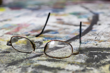 Etwas ramponierte Brille eines Künstlers liegt aus farbenfrohem Tisch in seinem Atelier